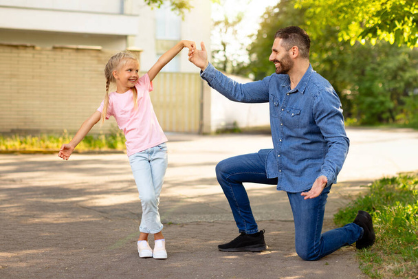 Kochający ojciec tańczący ze swoją uroczą córką na świeżym powietrzu w parku, szczęśliwy tata bawiący się i wygłupiający z dzieckiem, stojący na jednym kolanie i uśmiechnięty, przestrzeń do kopiowania - Zdjęcie, obraz