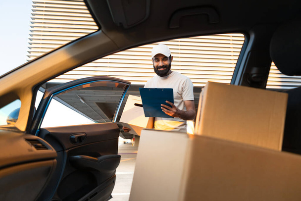 Delivering Orders On Time. Middle Eastern Delivery Guy Looking At Parcel Boxes In Car And Taking Notes Standing With Clipboard Near Auto, Wearing Beige Cap And T-Shirt Uniform. Selective Focus - Photo, Image