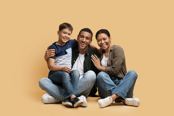 Happy cute loving family parents and child sitting on floor over colorful background, embracing and smiling at camera. Cheerful father, mother and schooler son enjoying time together - Photo, Image
