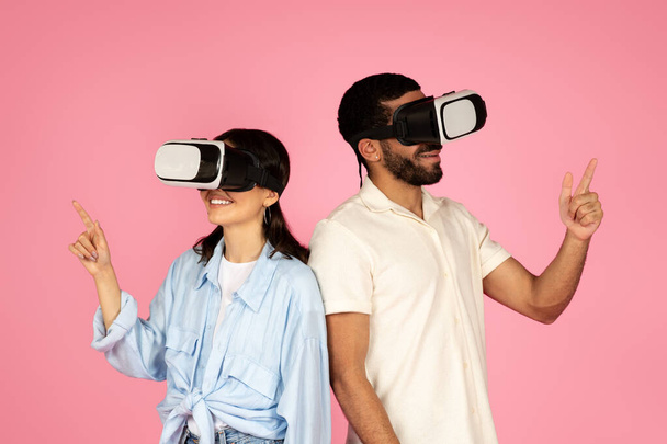 Jeu vidéo, simulations. Jeune homme et femme souriants en tenue décontractée profitant d'une expérience de réalité virtuelle, couple utilisant casque VR lunettes, toucher l'espace vide, isolé sur fond rose - Photo, image