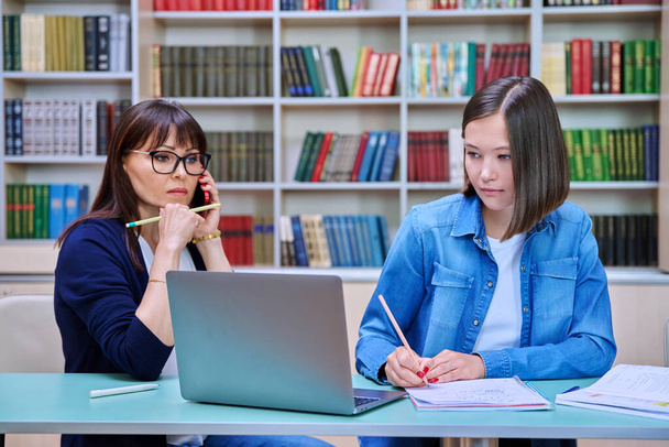 Young female university student with middle aged teacher mentor preparing for exam sitting at desk with laptop books inside library. Knowledge, education, youth, college university concept - Zdjęcie, obraz