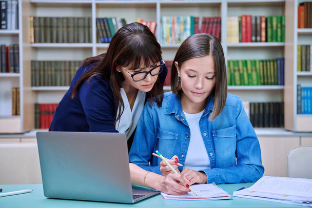 Young female university student with middle aged teacher mentor preparing for exam sitting at desk with laptop books inside library. Knowledge, education, youth, college university concept - Foto, imagen