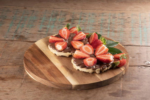 sobremesa de dois waffles com chocolate e creme de avelã em uma tábua de madeira redonda e morangos de redução em uma mesa de madeira - Foto, Imagem