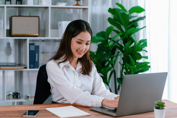 Mladá asijská nadšená podnikatelka na moderním kancelářském stole pomocí notebooku pracovat a psát poznámky. Pečlivá a atraktivní kancelářská dáma pracující na notebooku ve svém kancelářském prostoru. - Fotografie, Obrázek