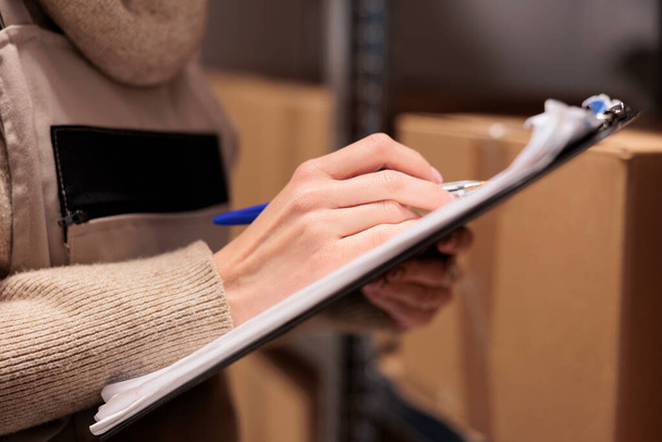 Postal office storage worker holding clipboard in warehouse, marking inventory checklist. Warehouse employee hand writing with pen close up, checking cardboard boxes in storehouse - Photo, Image