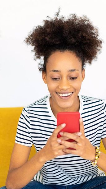 Genç Afrikalı bir kadının evdeki kanepede otururken akıllı telefon kullandığı dikey bir fotoğraf. Teknoloji ve sosyal medya kavramı. - Fotoğraf, Görsel