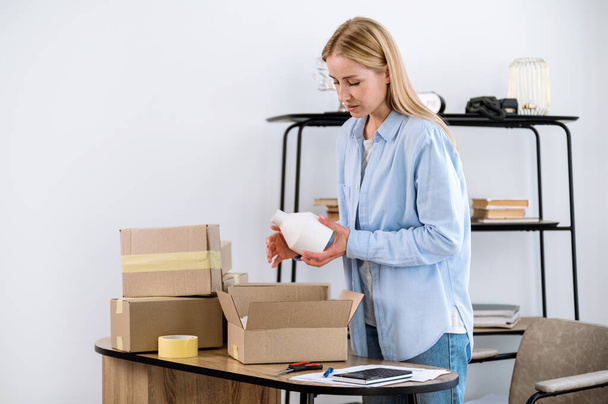 Female warehouse worker or salesperson packing fragile ceramic vase in cardboard box standing in storage room. Ecommerce shipping, delivery, small business and logistic service advertising concept. - Photo, Image