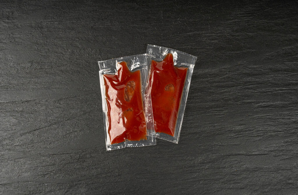 Ketchup en bolsa de plástico cuadrada en negro, bolsita de catsup transparente de una sola vez, salsa de tomate caliente, aderezo rojo, porción de ketchup en fondo oscuro - Foto, imagen