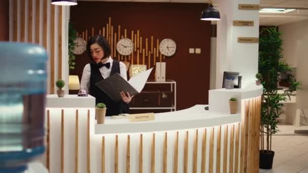 Happy friendly receptionist making phone calls standing at check in counter in welcoming resort lobby. Cheerful professional hotel employee taking booking requests while working at reception desk - Footage, Video