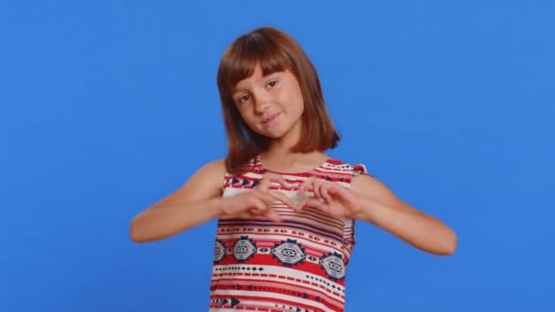 I love you. Smiling young cute brunette school girl makes heart gesture demonstrates love sign expresses good feelings and sympathy. Happy preteen female child kid isolated on studio blue background - Footage, Video