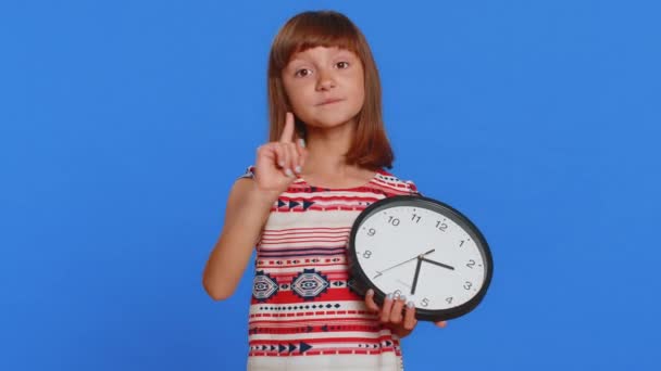 It is your time. Young school girl showing time on wall office clock, ok, thumb up, approve, pointing finger at camera, time for education study. Preteen child kid isolated on studio blue background - Footage, Video