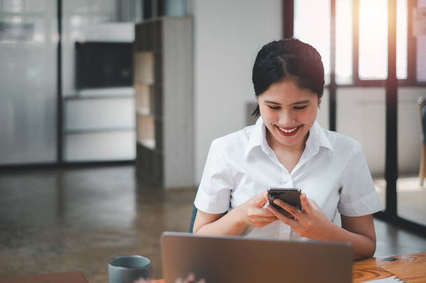 Attraente giovane donna che lavora sul cellulare e sorride mentre siede al suo posto di lavoro in ufficio e guarda il computer portatile - Foto, immagini