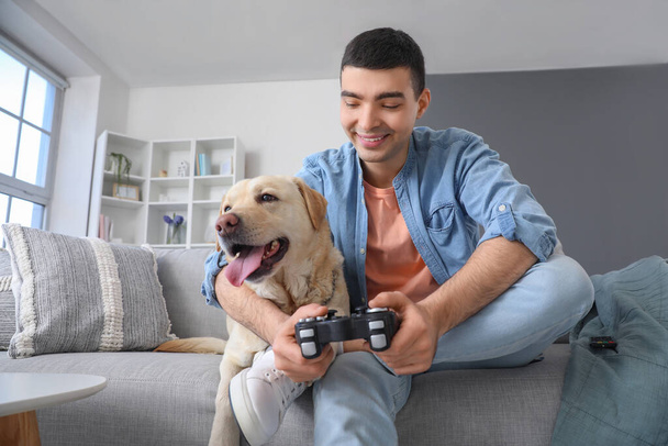 Νεαρός με χαριτωμένο σκυλί Λαμπραντόρ παίζει βιντεοπαιχνίδι στο σπίτι - Φωτογραφία, εικόνα
