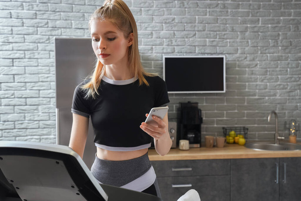 Sporty young woman with mobile phone training on treadmill at home - Photo, Image