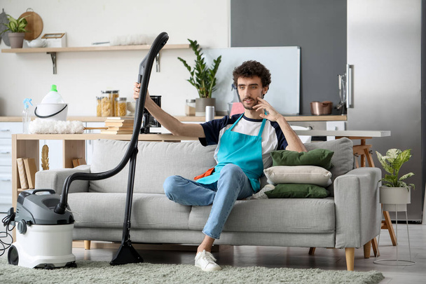 Young man with vacuum cleaner sitting on sofa at home - Foto, afbeelding