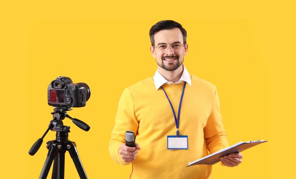 Male journalist with microphone and clipboard recording video on yellow background - Photo, Image