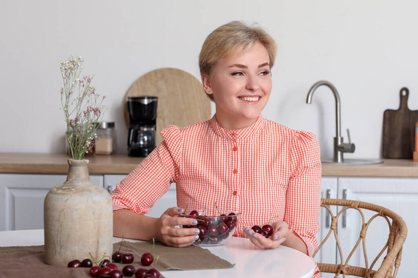Beautiful young happy woman with bowl of ripe cherries sitting at table in kitchen - Fotoğraf, Görsel
