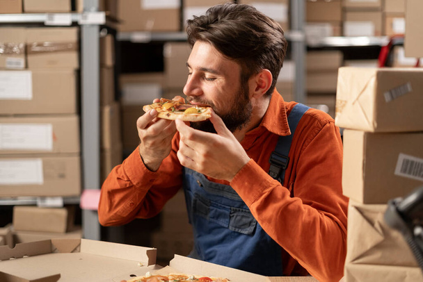 Jonge magazijnmedewerker die pizza eet tijdens de lunch op het werk, voedselconcept op het werk. Close-up - Foto, afbeelding
