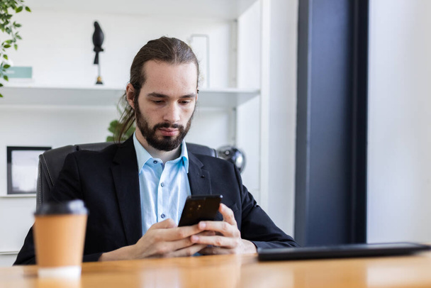 Young caucasian handsome businessman with beard using smartphone sitting office table. Personal mobile communication. Man send message or search browsing information. Ordering work via online system. - Photo, image