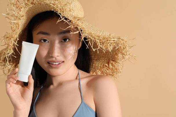 Beautiful young woman in straw hat with sunscreen on her face holding sun protection cream against beige background, space for text - Фото, зображення
