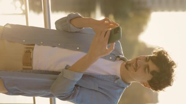 Vertikales Video. Junger Mann, der mit seinem Smartphone auf der Straße steht und mit Freunden SMS schreibt. Kaukasisch lächelnder Mann draußen scrollt unterwegs mobil. Teenager chatten im Internet - Filmmaterial, Video