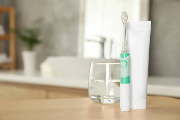 Electric toothbrush, tube with paste and glass of water on wooden table in bathroom. Space for text - Фото, изображение