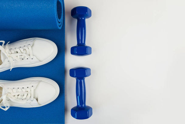 Fitness, yoga, spor, sağlıklı yaşam tarzı. Beyaz spor ayakkabıları, mavi halterler ve beyaz arka planda mavi egzersiz paspası. Boşluğu kopyala. - Fotoğraf, Görsel