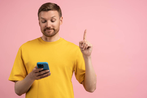 Kiinnostunut mies etsii älypuhelimen näytön ottaa oivallus osoittaa sormella ylös eristetty vaaleanpunainen studio seinään. Eurekaa. Tyytyväinen kaveri löysi tarvittavat tiedot matkapuhelin poseeraa taustakopiona tilaa - Valokuva, kuva