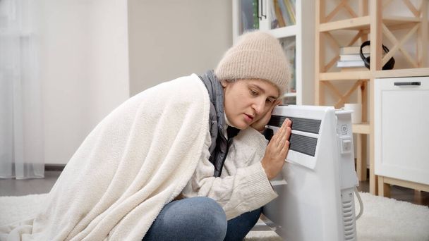 Młoda kobieta w zimowych ubraniach siedzi w domu i grzeje się w ogrzewaczu elektrycznym. - Zdjęcie, obraz