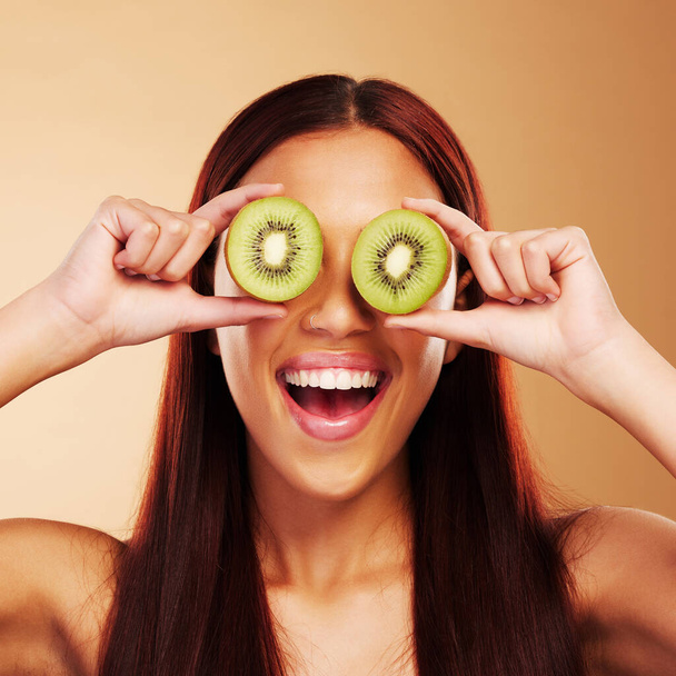 Beauty, kiwi and a woman with skin care in studio for natural dermatology, cosmetics or glow. Facial, fruit and healthy diet for vitamin c and nutrition of excited model person on a brown background. - Photo, Image