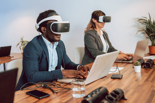 Konzentrierter afroamerikanischer Unternehmer mit VR-Brille sitzt mit Laptop am Tisch und arbeitet im Büro an einem Projekt - Foto, Bild