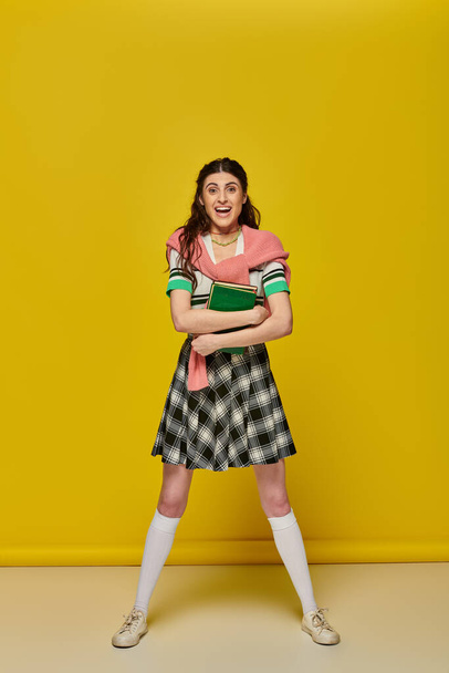 excited young woman in skirt standing with books on yellow backdrop, happy student, college outfit - Photo, Image