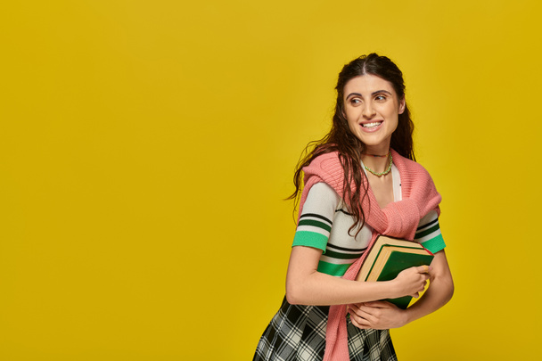 позитивная молодая женщина в юбке стоя с книгами на желтом фоне, счастливый студент, колледж наряд - Фото, изображение