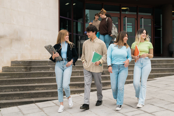 Full length group of positive young male and female students with notebooks in hands smiling and chatting while walking on street after university - Photo, Image