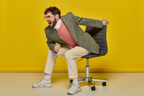 érzelmi és szakállas férfi sikoltozik és ül irodai szék, sárga háttér, dühös diák - Fotó, kép