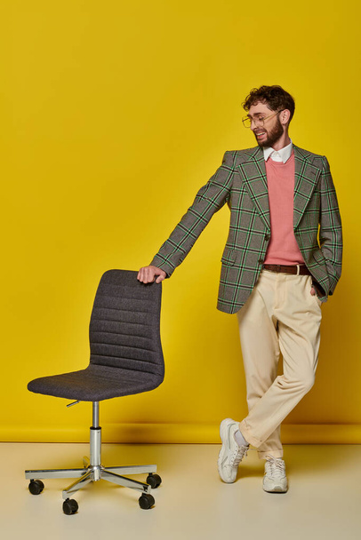 hombre positivo de pie con la mano en el bolsillo cerca de la silla de oficina, fondo amarillo, estudiante en gafas - Foto, imagen