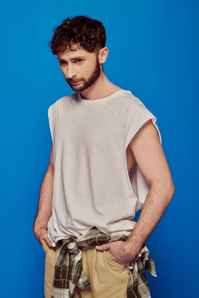 bearded and curly man standing with hands in pockets on blue background, tank top, male fashion - Фото, зображення