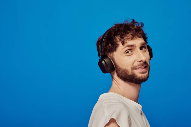 pozytywny mężczyzna w słuchawkach bezprzewodowych słuchanie muzyki na niebieskim tle, moda męska, audio - Zdjęcie, obraz