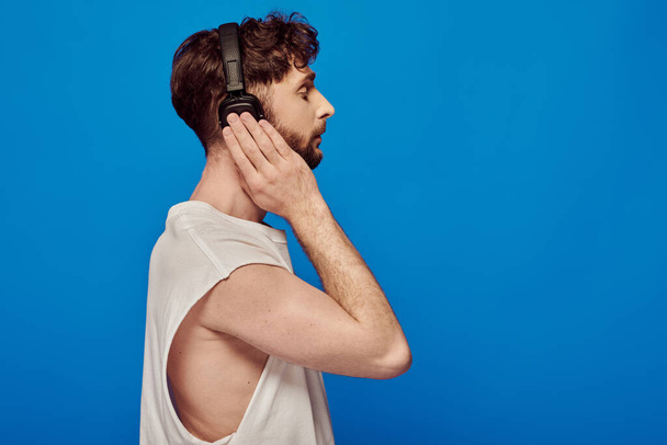 side view, bearded man in wireless headphones listening music on blue background, summer fashion - Photo, Image