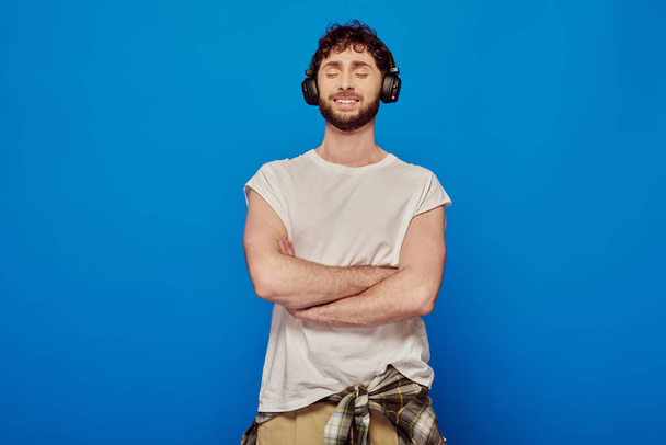 θετικός άνδρας σε ασύρματα ακουστικά ακούγοντας μουσική σε μπλε φόντο, κλειστά μάτια, καλοκαιρινή μόδα - Φωτογραφία, εικόνα