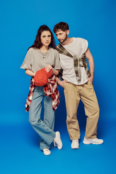 college couple posing in street wear on blue backdrop, woman with bold makeup, baseball cap - Photo, Image