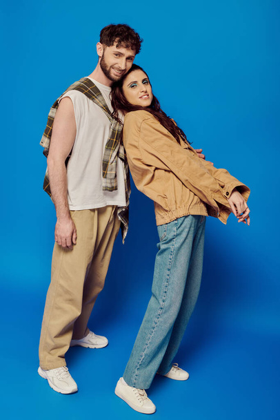happy and stylish couple in casual wear posing on blue backdrop, woman and man looking at camera - Foto, Imagem