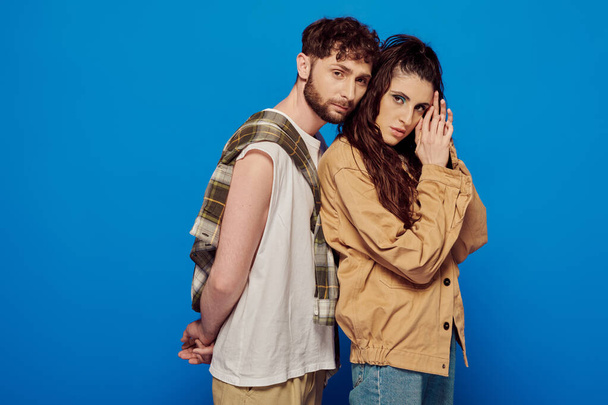 stylish couple in casual wear posing on blue backdrop, woman and man with beard looking at camera - Zdjęcie, obraz