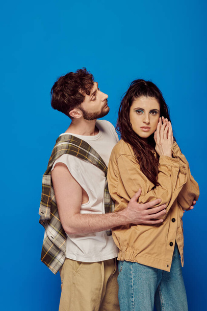 stylish couple in casual wear posing on blue backdrop, bearded man hugging woman, looking at camera - Photo, Image