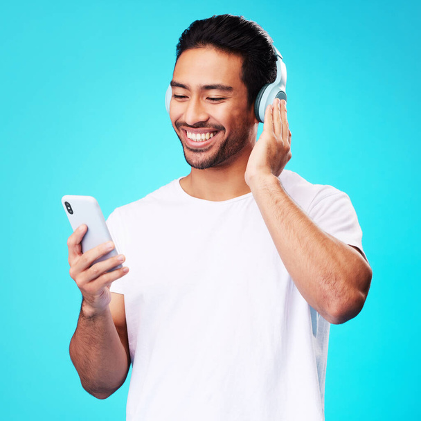 Fejhallgató, zene és ázsiai férfi okostelefonnal a stúdióban való hallgatáshoz elszigetelt kék háttérrel. Rádió, mosoly és személy mobil, hang vagy hallás podcast, audio vagy közösségi média az interneten. - Fotó, kép