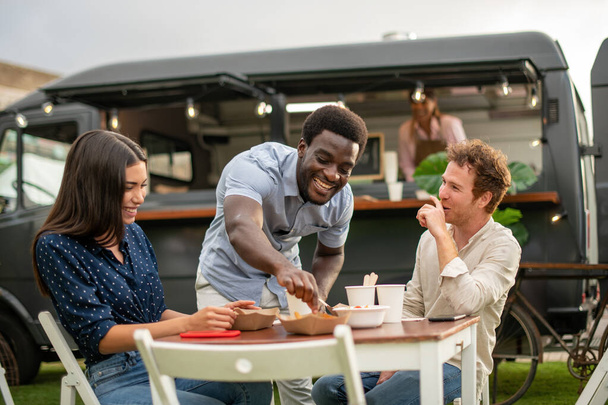 Company of positive multiethnic friends gathering at table and eating takeaway fast food in park with food truck - Photo, Image