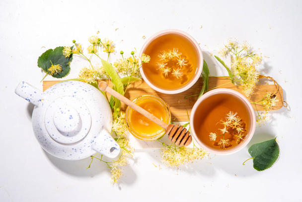 Organik bitkisel sağlık içeceği. Linden çiçeği yeşili çay, beyaz kupalar, çaydanlık, beyaz bir masa, kopya alanı ve çiçek açan ıhlamur çiçekleri - Fotoğraf, Görsel