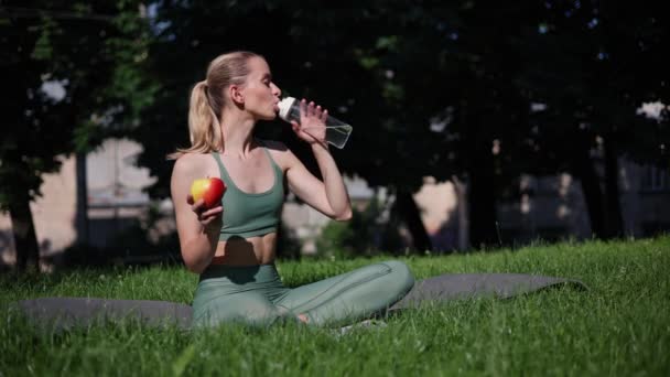 Fitness kızı nilüfer çiçeği pozunda oturur, spor şişesinden su yudumlar ve elma yer. - Video, Çekim