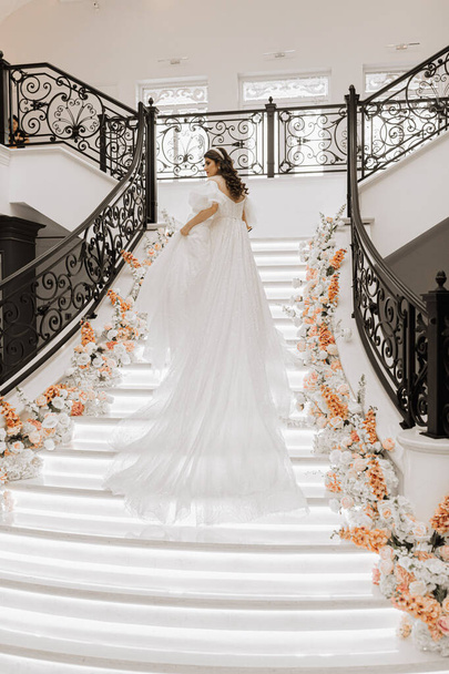 full-length portrait of a young girl from the back, with beautiful hair and makeup in a luxurious wedding dress on the steps of a restaurant near rose flowers. Vertical photo. - 写真・画像