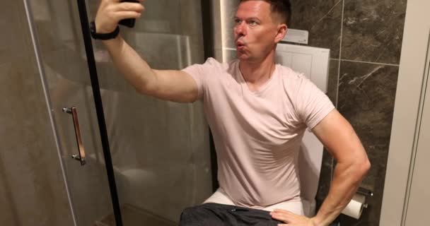 Homme heureux prend selfie photo assis dans la salle de bain sur les toilettes. Guy grimace et prend des photos - Séquence, vidéo